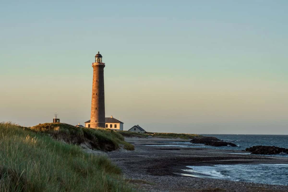 Phare de brique rouge dressé au-dessus d'une dune herbeuse, au bord d'une côte de galets, dans la lumière du soir