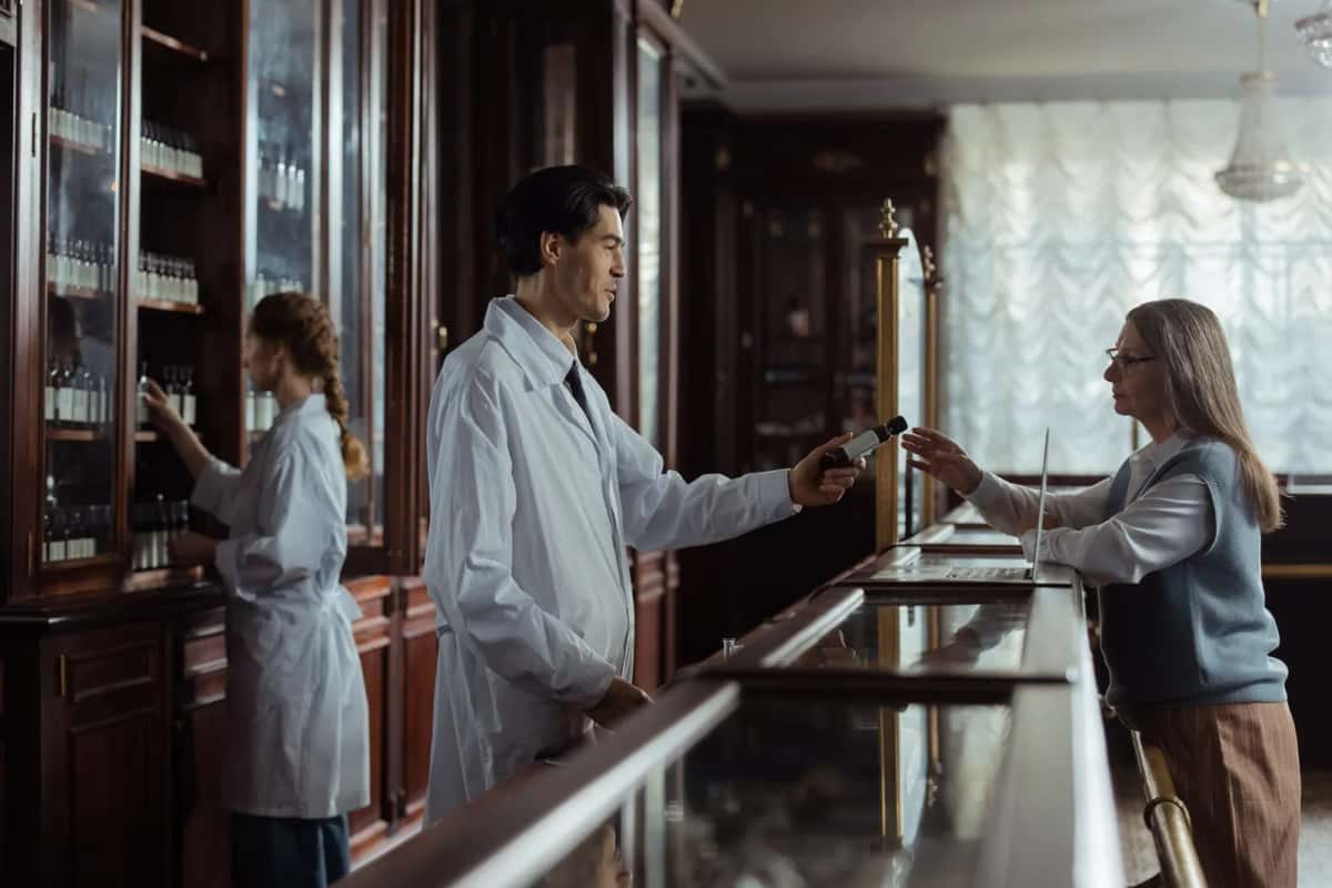 Comptoir de pharmacie aux boiseries anciennes, un préparateur en blouse remet une boîte à une cliente