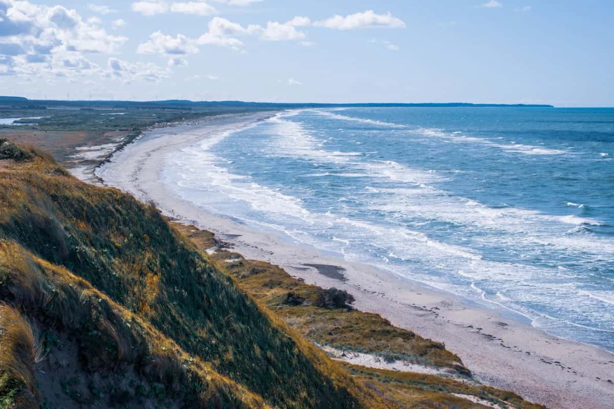 Longue plage de sable bordée de dunes couvertes d'oyats, vagues déferlantes et éoliennes à l'horizon