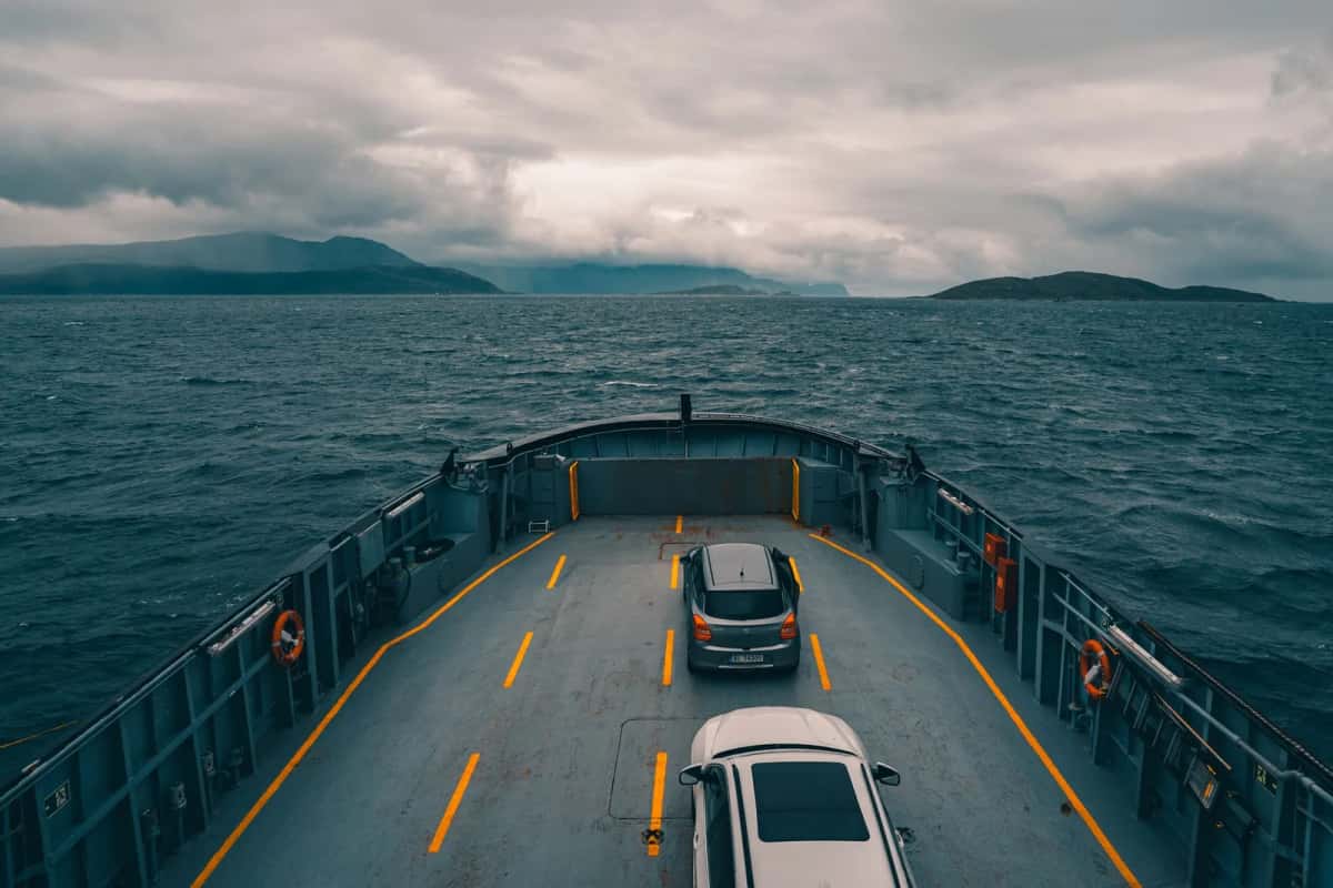 Pont-garage d'un ferry ouvert sur la mer, deux voitures garées entre les marquages jaunes, côte montagneuse à l'horizon