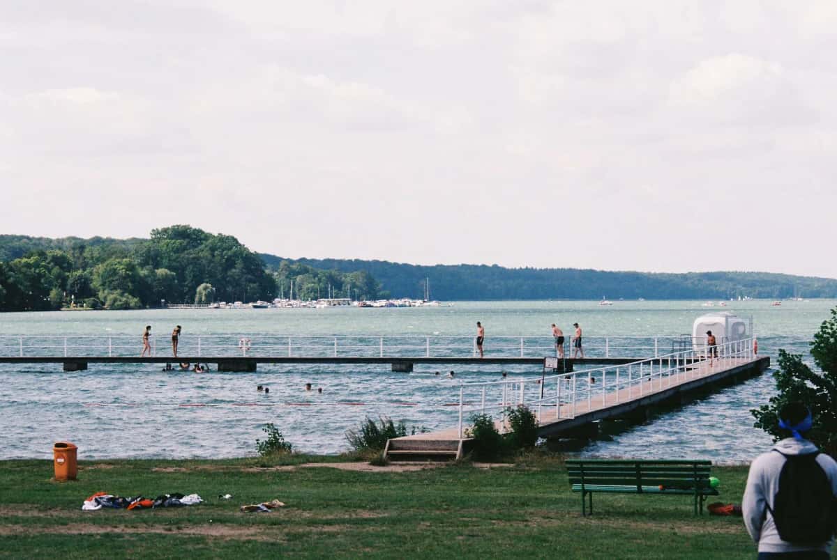 Ponton de baignade en bois s'avançant sur l'eau, quelques nageurs et une pelouse bordée de bancs au premier plan