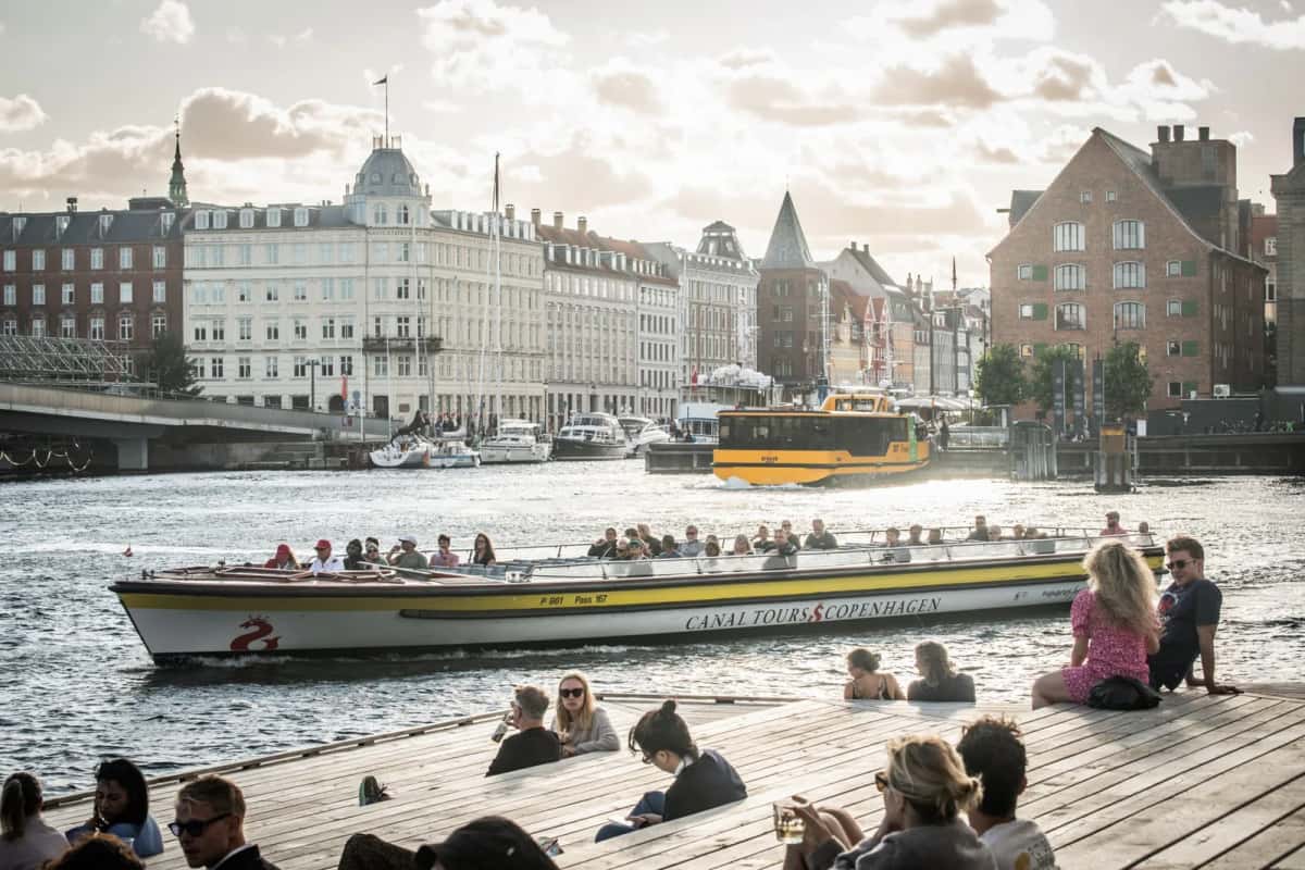 Des gens assis sur un quai en bois au bord d'un canal de Copenhague, un bateau de promenade Canal Tours passant devant eux en fin de journée