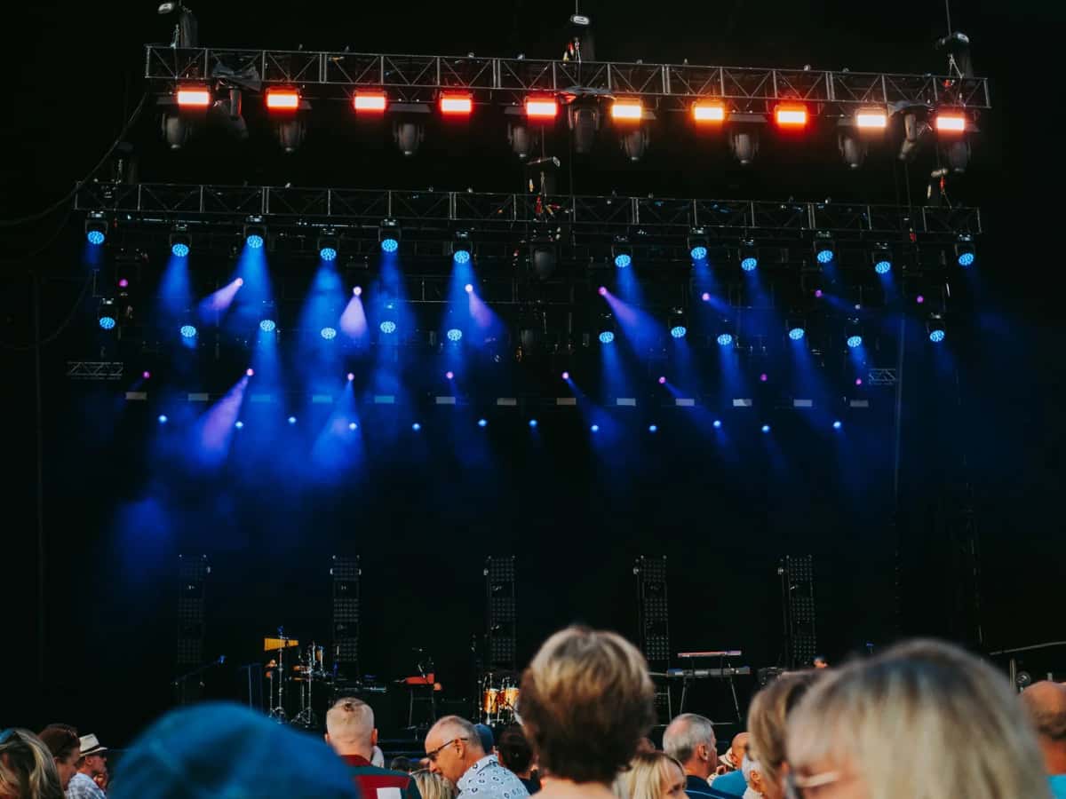 Rampe de projecteurs bleus et rouges suspendue au-dessus d'une scène de plein air, quelques spectateurs de dos au premier plan