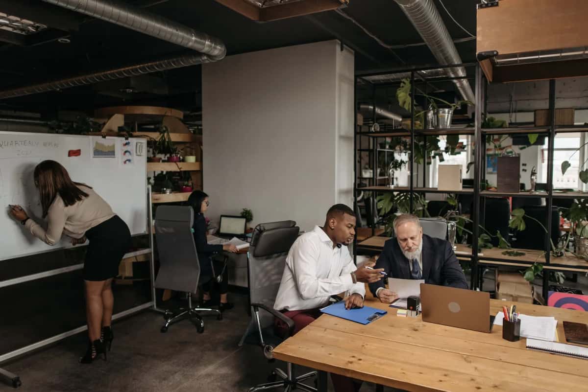 Réunion d'équipe autour d'une grande table de bureau dans un plateau ouvert, ordinateurs portables et tableau blanc en arrière-plan
