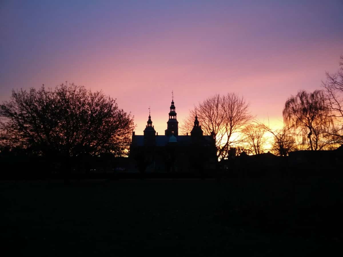 Silhouette des tourelles et pignons du château de Rosenborg se découpant sur un ciel rose et orange au coucher du soleil, arbres nus au premier plan