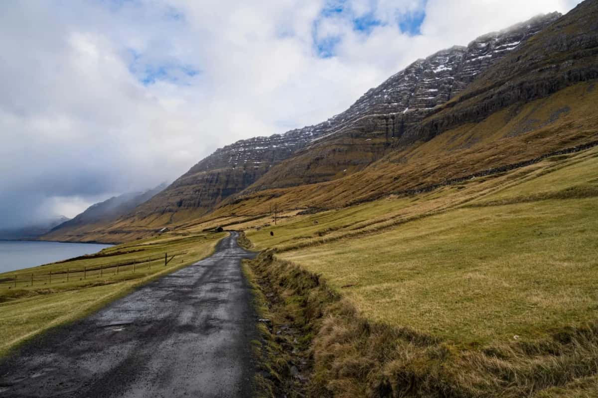 Route à une voie serpentant entre une pente herbeuse et un bras de mer, sous une montagne encore mouchetée de neige