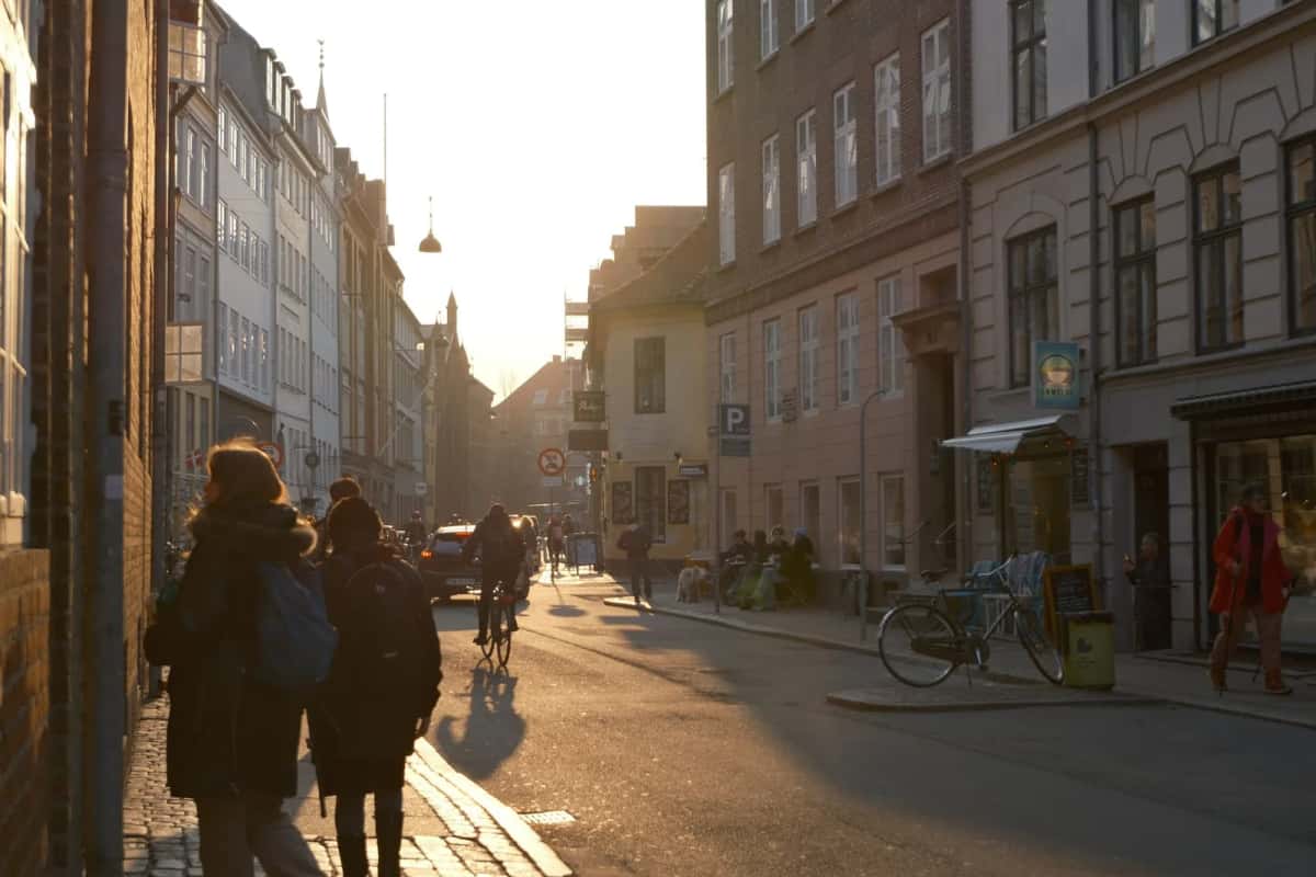 Rue étroite bordée d'immeubles anciens, des passants et des cyclistes à contre-jour dans la lumière rasante de fin de journée