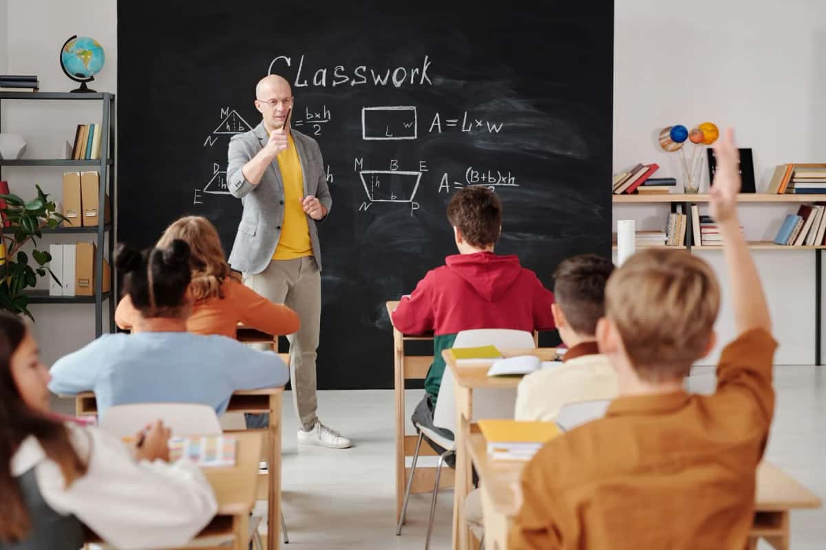Un enseignant debout devant un tableau noir couvert de formules, face à une classe d'adultes et d'adolescents assis, une main levée