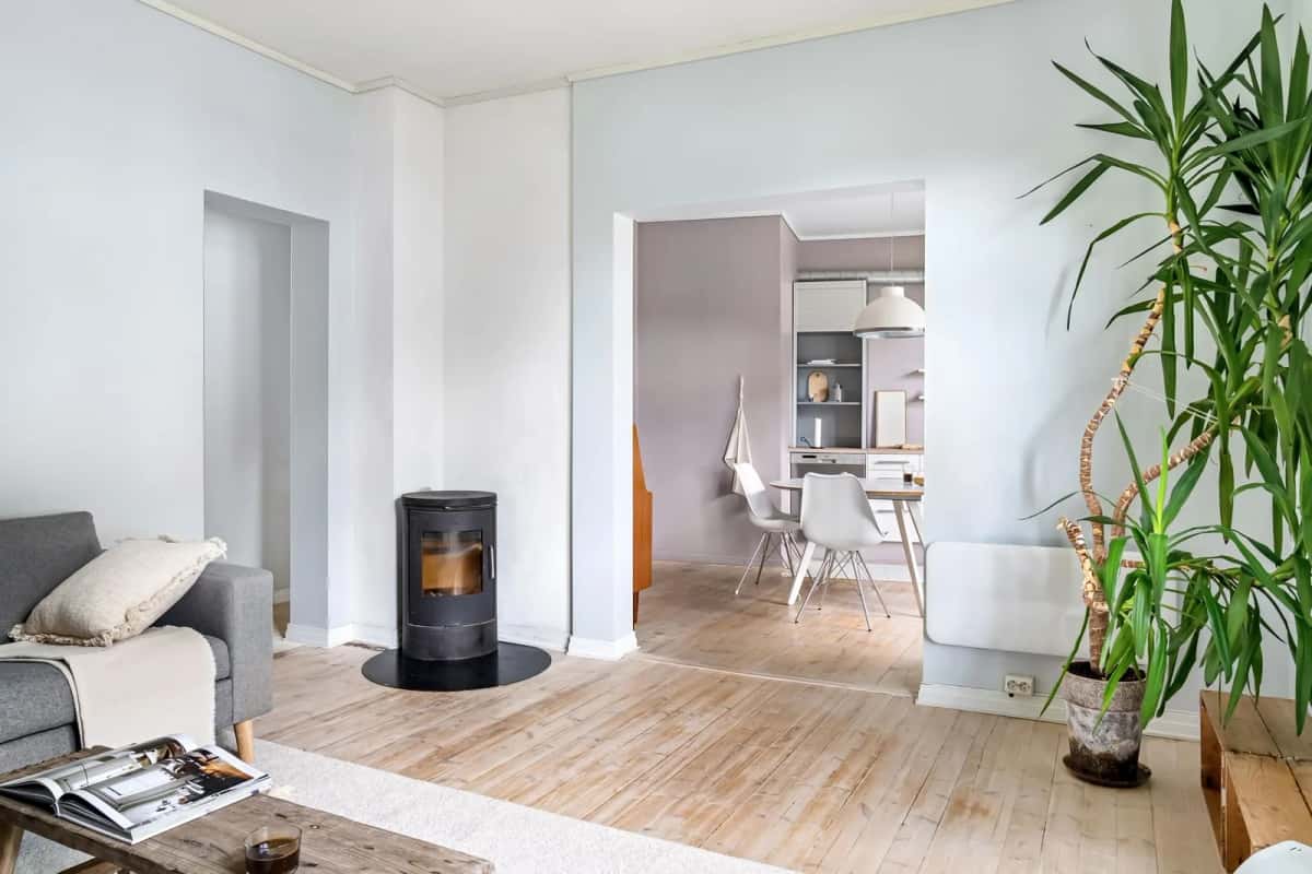 Salon nordique aux murs blancs et au parquet clair, poêle à bois noir allumé, canapé bas et grandes plantes vertes