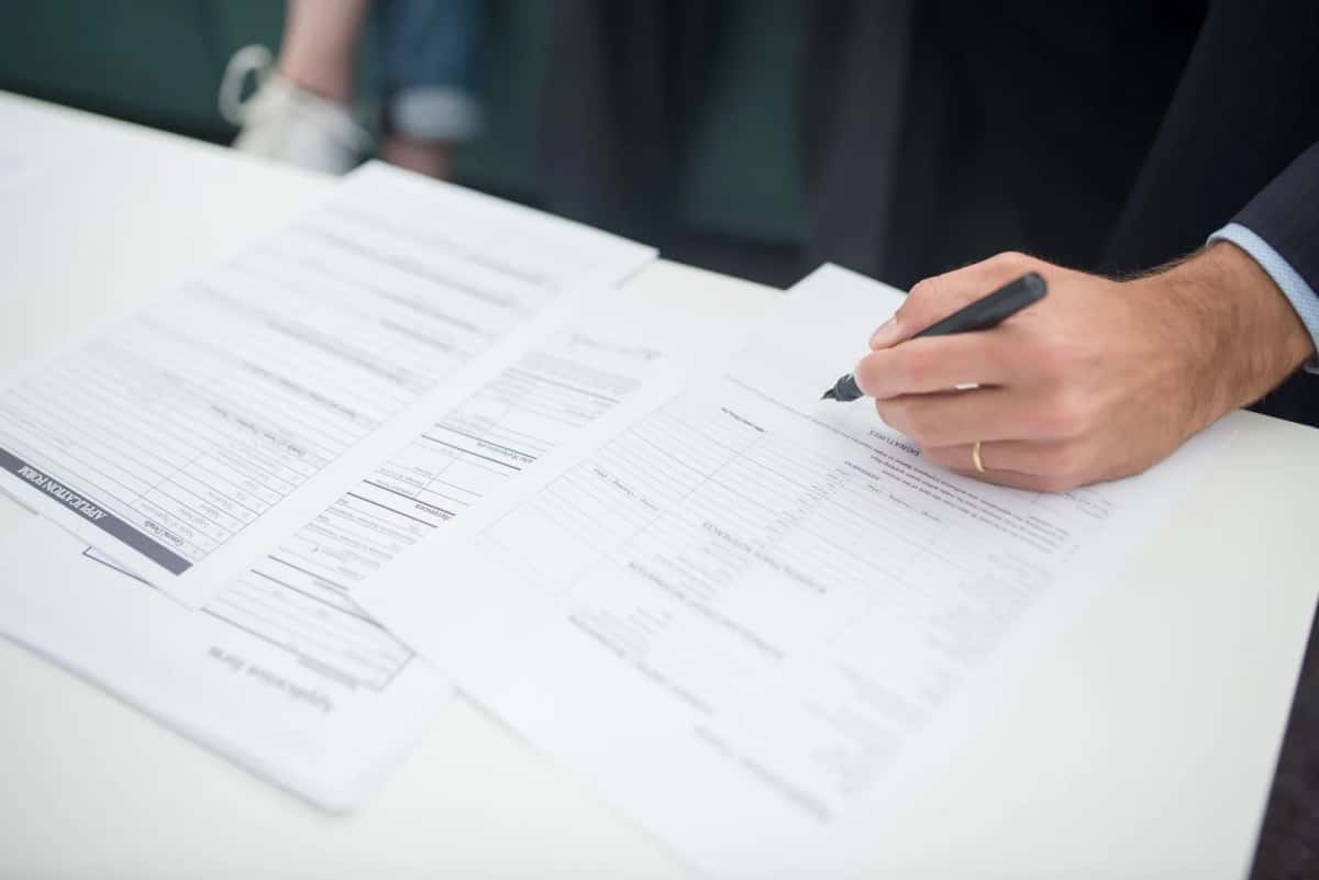 Une main signant un formulaire administratif posé sur une table blanche, à côté d'un second document