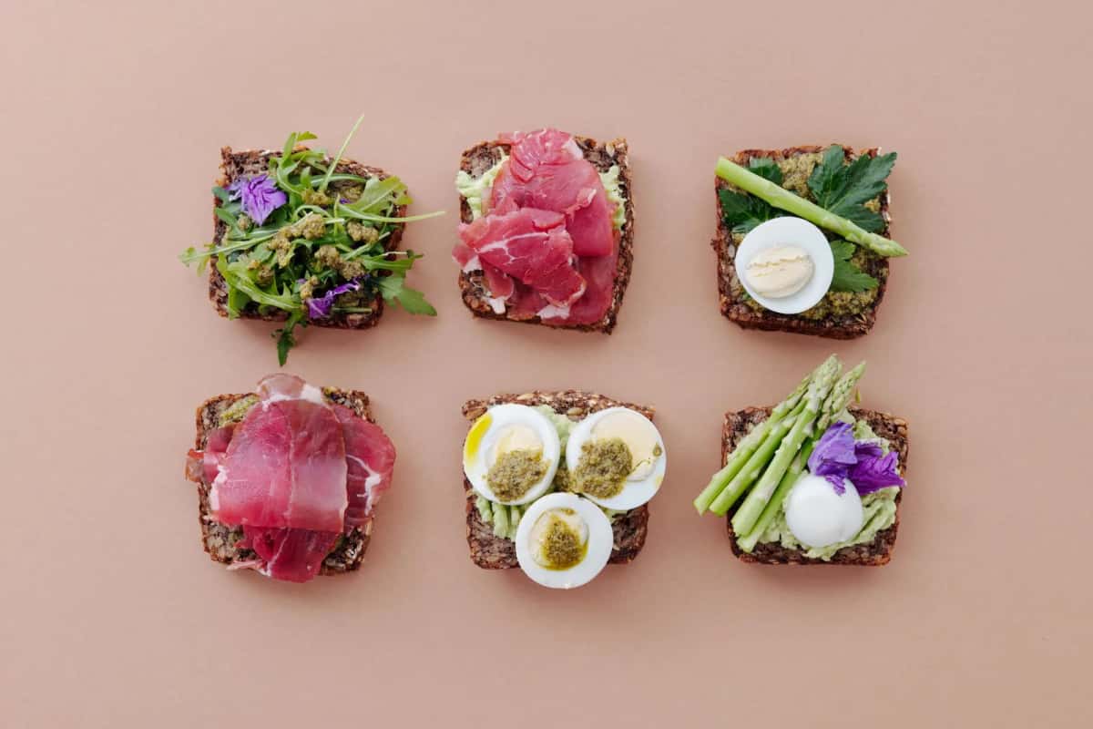 Six tartines de pain de seigle garnies de rosbif, d'œuf dur, d'asperges vertes et de pousses, disposées sur un fond rose
