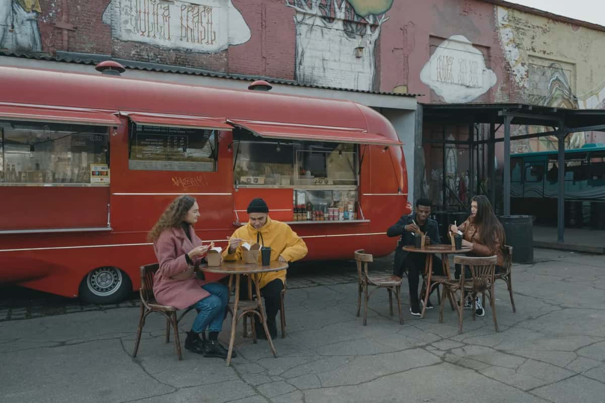 Caravane rouge transformée en comptoir de street food, quelques tables et tabourets en extérieur devant des murs couverts de graffitis