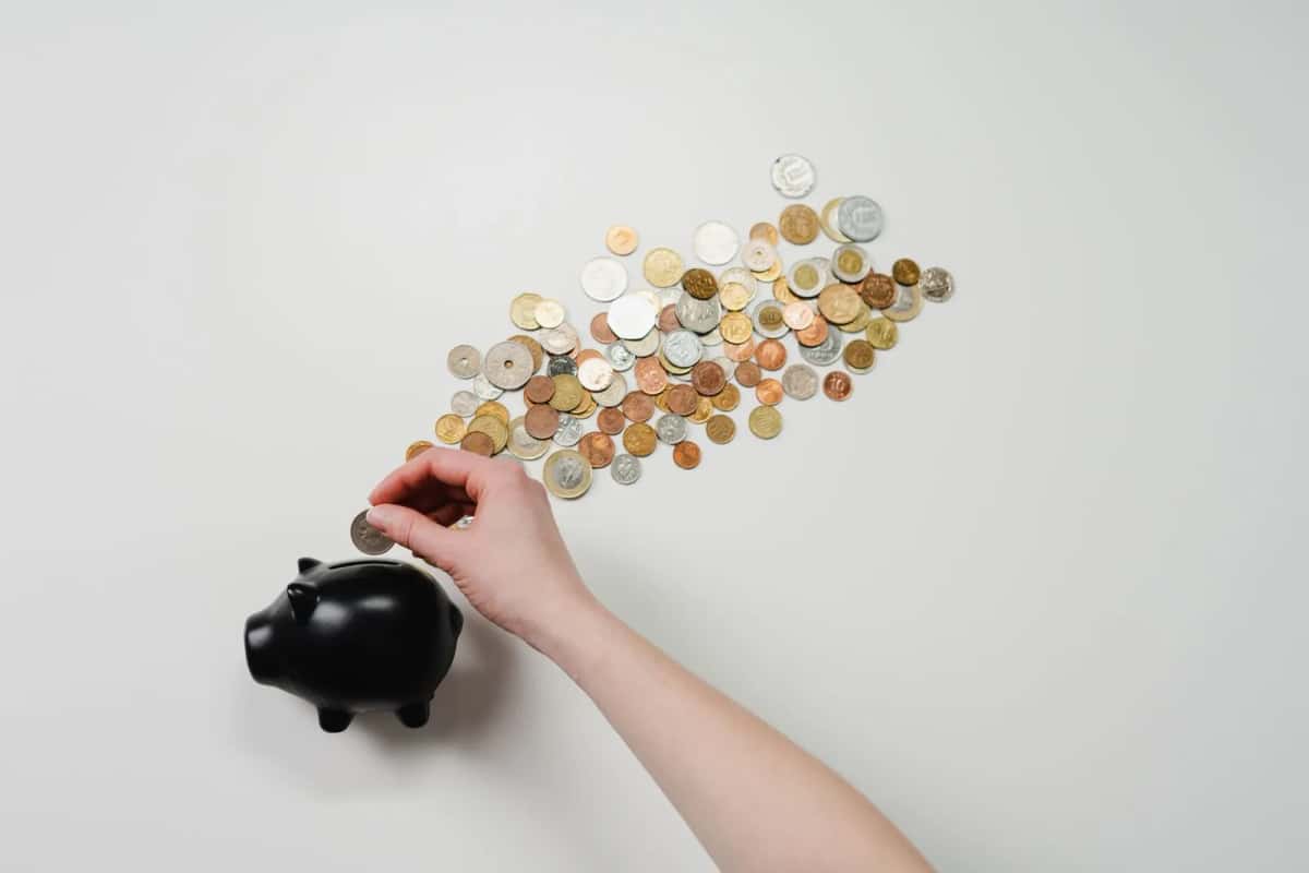 Une main glissant une pièce dans une tirelire noire, à côté d'un large éventail de pièces de monnaie étalées
