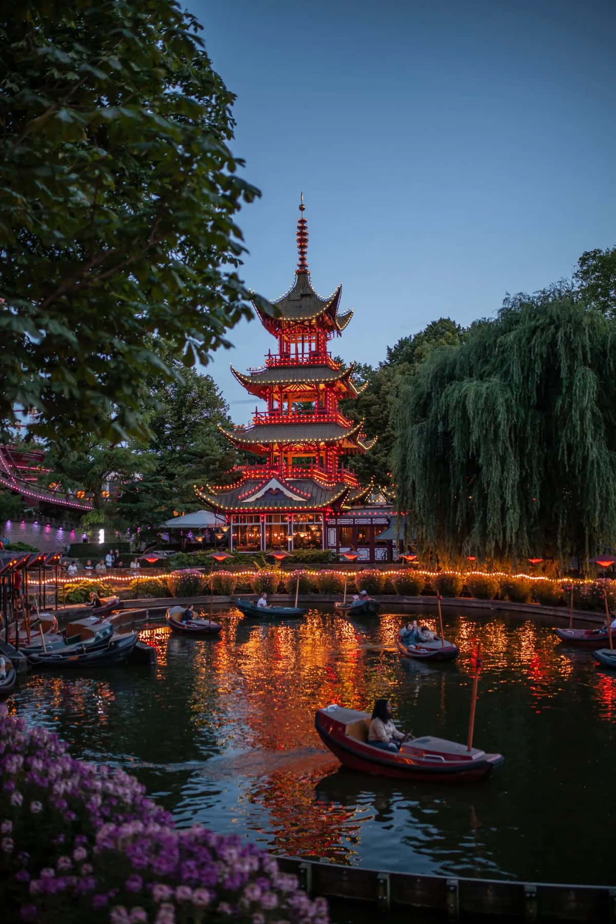 La pagode chinoise illuminée des jardins de Tivoli à Copenhague à l'heure bleue, reflets orange sur le lac et barques à moteur menées par des visiteurs