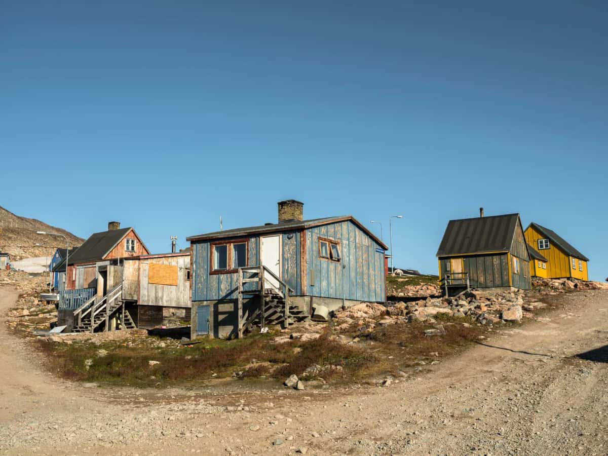 Maisons de bois bleues, jaunes et grises posées sur pilotis le long d'une piste de gravier, dans un village arctique sans arbres